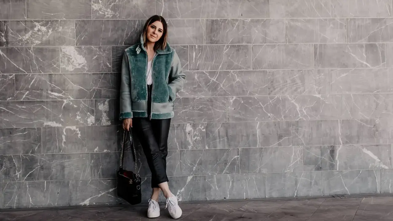 Frau in mintgrüner Lammfelljacke und Lederhose, die weiße Sneakers trägt. Sie steht vor einer grauen Marmorwand.