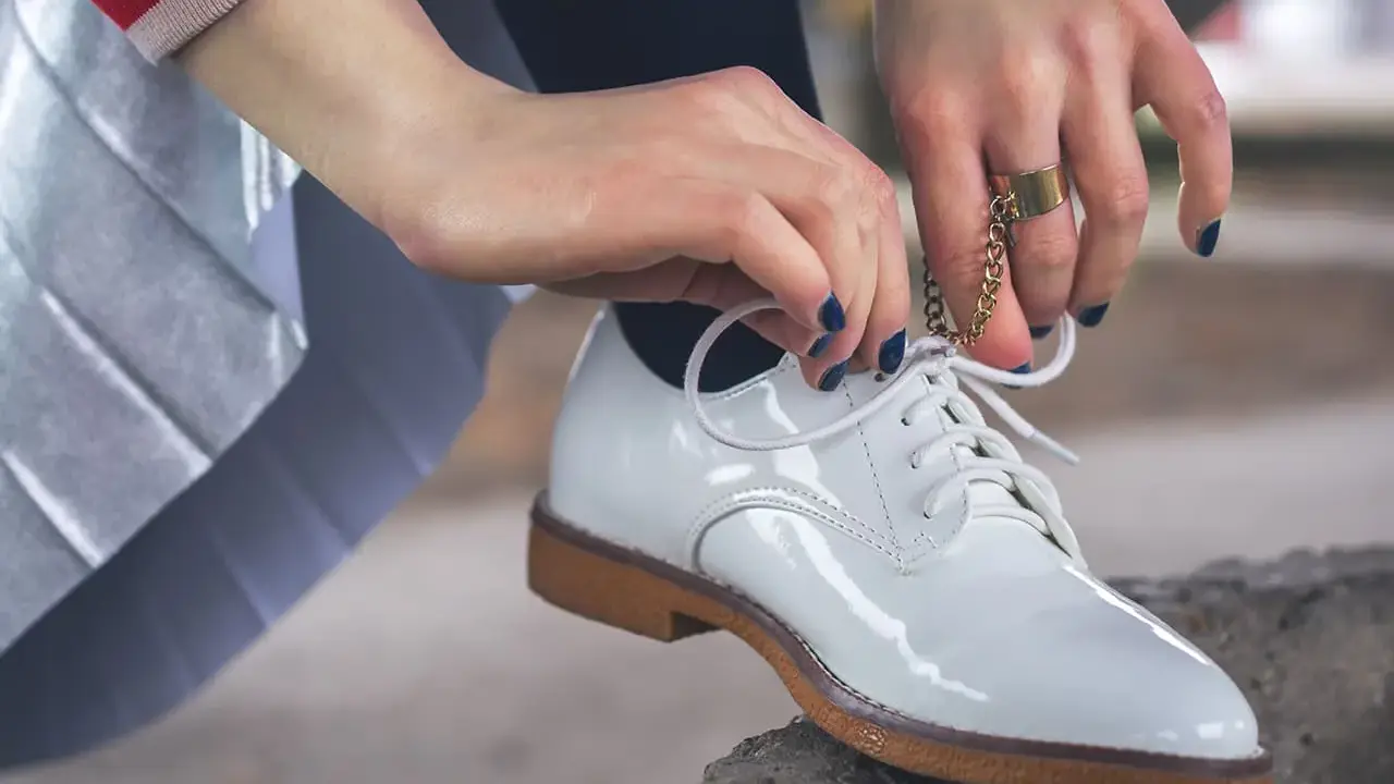 Hände mit dunkelblauem Nagellack binden weiße Lackschuhe. Die Pflege von Lackschuhen ist wichtig, damit sie glänzen.
