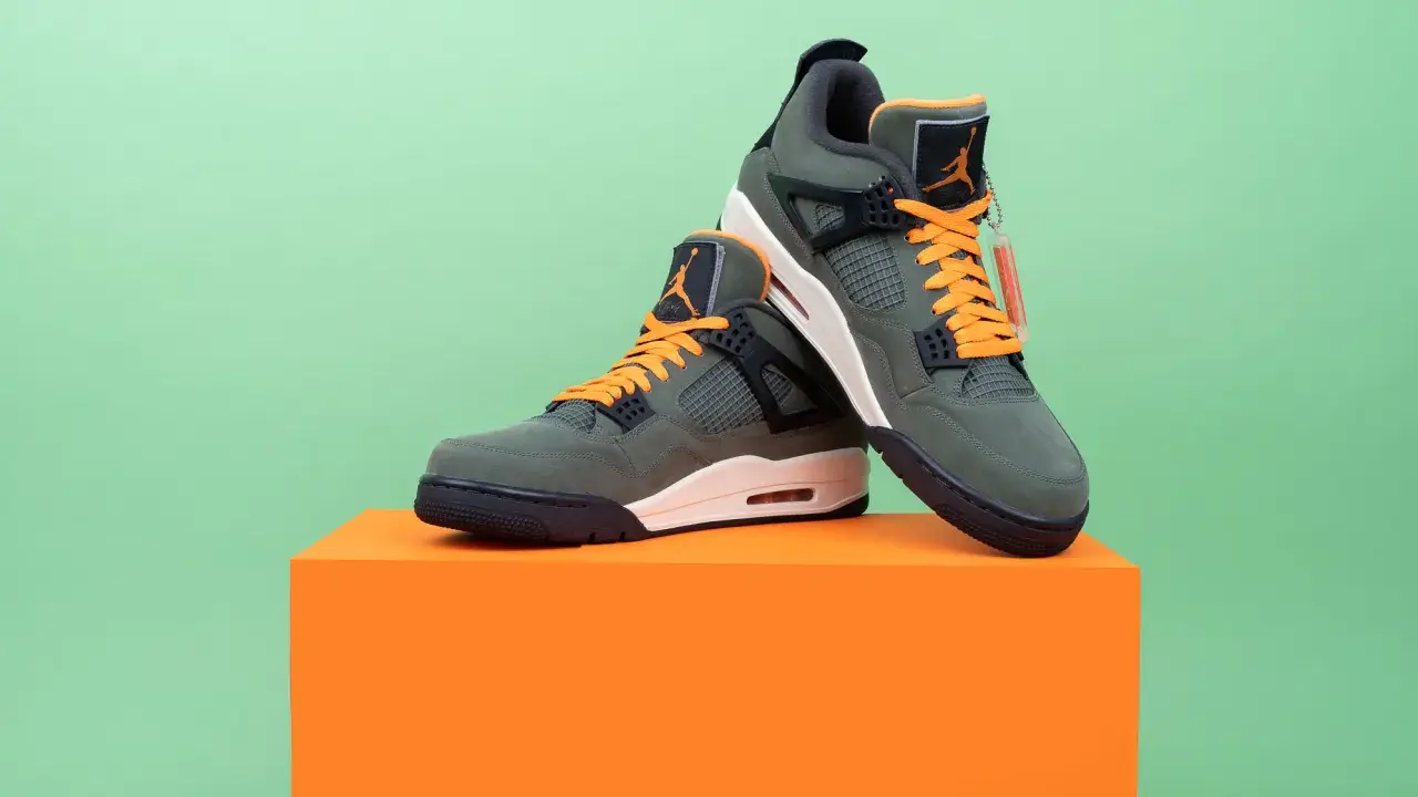 Jordan Motorsport Sneaker in Olivgrün mit orangefarbenen Schnürsenkeln, auf einem orangefarbenen Podest vor hellgrünem Hintergrund.