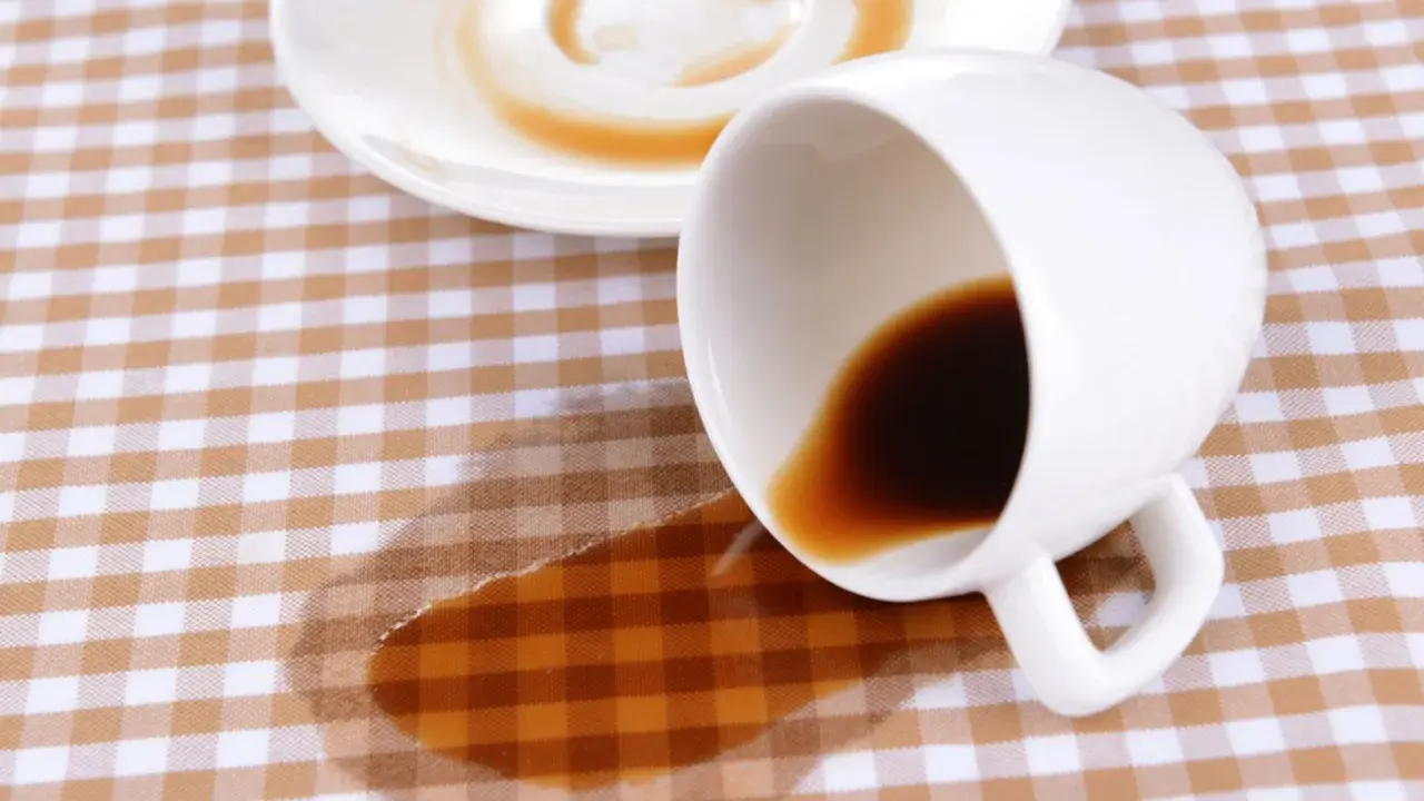 Um Kaffeeflecken zu entfernen, ist hier ein umgekippter Becher mit Kaffee auf kariertem Stoff.