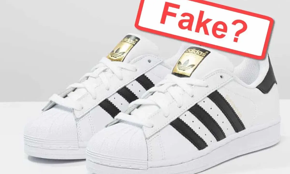 Weiße Adidas Sneaker mit schwarzen Streifen und goldenem Logo. Ein rotes Schild fragt: "Fake?". So kannst du Adidas Fake erkennen.