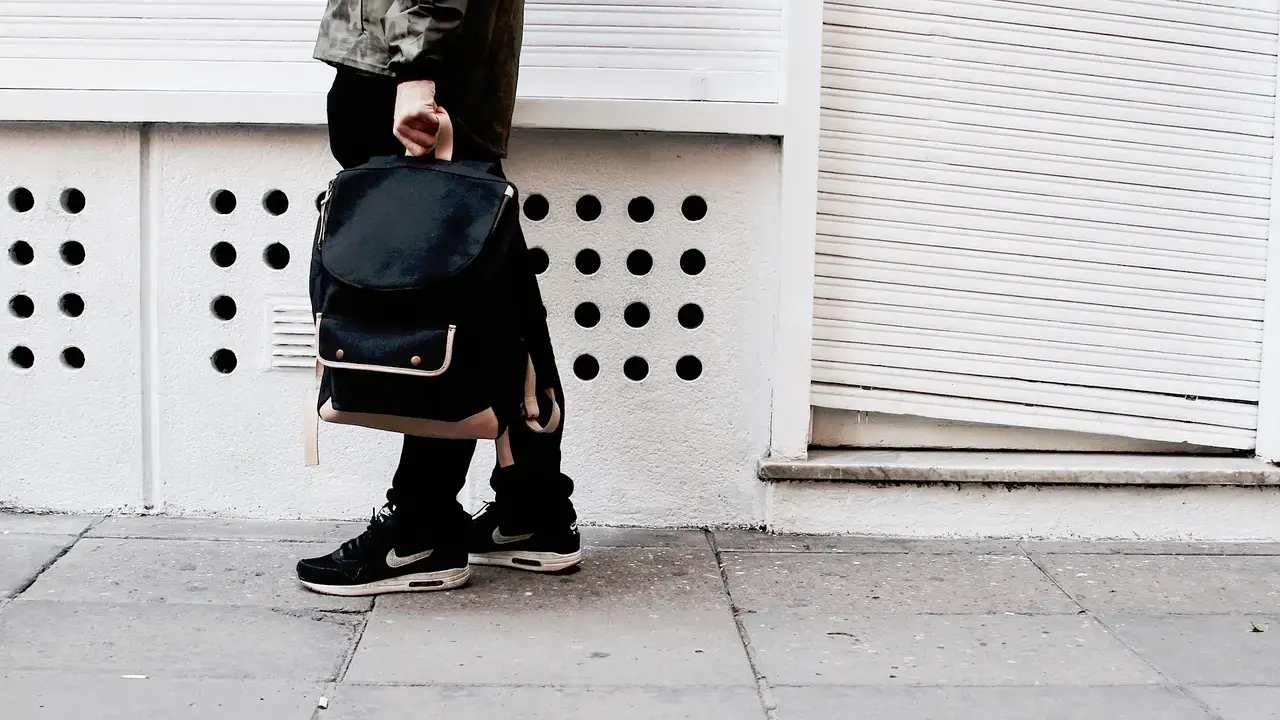 Person mit schwarzem Rucksack und **air max fake** Schuhen steht auf einem Gehweg vor einer weißen Wand mit runden Öffnungen.