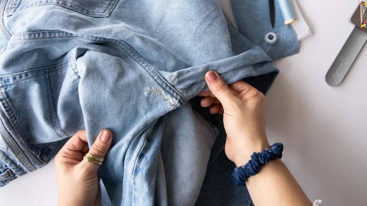 Hände halten hellblaue Jeans, die zum Nähen vorbereitet werden. Nähutensilien wie Schere und Garn liegen bereit. Vielleicht werden die Jeans bald ins Gefrierfach gelegt, um sie zu erfrischen.