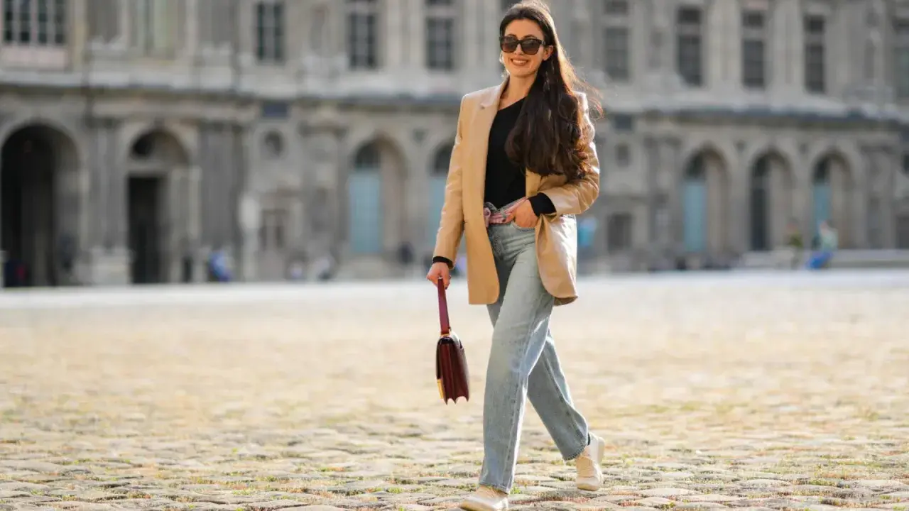 Eine Frau mit langen Haaren und Sonnenbrille spaziert lässig in weiten Jeans und einem beigen Blazer vor einer historischen Fassade.
