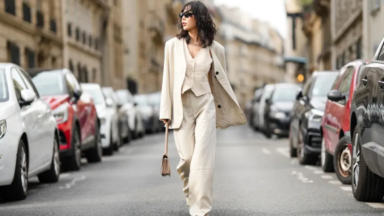 Eine Frau in einem eleganten beigen Hosenanzug mit passendem Westchen und Blazer spaziert durch eine Pariser Straße. Die weiten Hosen und die lässige Silhouette der Schuhe zu Hosenanzug Damen sind perfekt für einen stilvollen Auftritt.