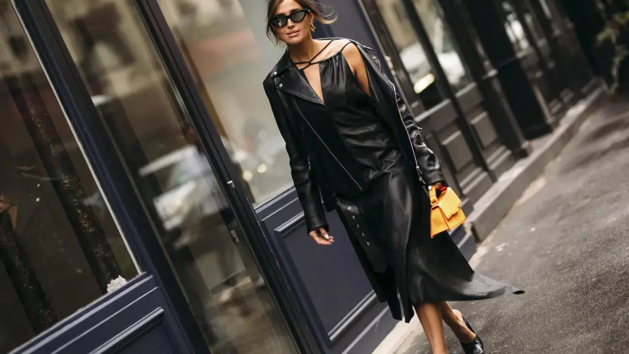 Stilvolle Frau in einem schwarzen Lederkleid und passender Lederjacke, die zeigt, wie man ein schwarzes Kleid kombinieren kann. Orange Handtasche als Farbtupfer.