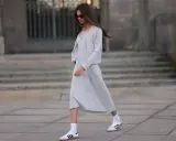 Frau in grauem Strick-Abendkleid mit Cardigan und weißen Sneakern, die lässig über einen Platz geht.