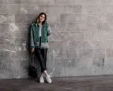 Frau in mintgrüner Lammfelljacke und Lederhose, die weiße Sneakers trägt. Sie steht vor einer grauen Marmorwand.