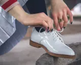Hände mit dunkelblauem Nagellack binden weiße Lackschuhe. Die Pflege von Lackschuhen ist wichtig, damit sie glänzen.