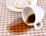 Um Kaffeeflecken zu entfernen, ist hier ein umgekippter Becher mit Kaffee auf kariertem Stoff.