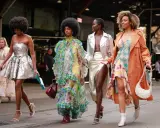 Vier Frauen in auffälligen Outfits. Eine trägt eine silberne Paillettenjacke zum Kleid, eine andere ein buntes Rüschenkleid.