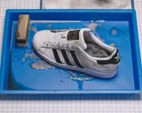 Ein schmutziger weißer Adidas Superstar Schuh liegt in einer blauen Wanne mit Wasser und Seife. Reinigungswerkzeuge liegen daneben.