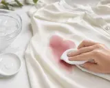 Hand tupft mit einem Tuch einen roten Weinfleck von einem weißen Hemd. Eiswasser und ein Tropfen Reinigungsmittel liegen bereit, um den Rotweinfleck zu entfernen.