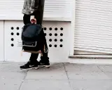 Person mit schwarzem Rucksack und **air max fake** Schuhen steht auf einem Gehweg vor einer weißen Wand mit runden Öffnungen.