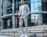 Ein Mann trägt einen hellgrauen Anzug mit weißem T-Shirt und Sneakern. Er steht auf einer Treppe vor einem modernen Gebäude.
