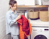 Frau prüft einen roten und orangen Daunenmantel, bereit zum wollmantel waschen. Daneben stehen Wäschekörbe und eine Waschmaschine.