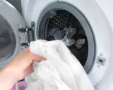 Eine Hand hält ein weißes Hemd, bereit, es in die Waschmaschine zu legen. Die Trommel ist bereits mit anderer Wäsche gefüllt.