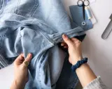 Hände halten hellblaue Jeans, die zum Nähen vorbereitet werden. Nähutensilien wie Schere und Garn liegen bereit. Vielleicht werden die Jeans bald ins Gefrierfach gelegt, um sie zu erfrischen.