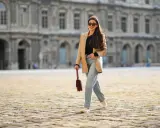 Eine Frau mit langen Haaren und Sonnenbrille spaziert lässig in weiten Jeans und einem beigen Blazer vor einer historischen Fassade.