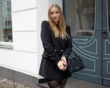 Frau mit blonden Haaren, die ein schwarzes Kleid mit Blazer kombiniert. Sie trägt eine schwarze Tasche und graue Stiefel.