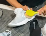 Hände reinigen weiße Mesh-Schuhe mit einer Bürste. Daneben liegen Schnürsenkel und eine Einlegesohle.