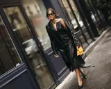Stilvolle Frau in einem schwarzen Lederkleid und passender Lederjacke, die zeigt, wie man ein schwarzes Kleid kombinieren kann. Orange Handtasche als Farbtupfer.