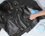 Schwarze Lederjacke reinigen mit einem feuchten Tuch. Eine Hand wischt über die Jacke, daneben eine Schale mit Seifenwasser.