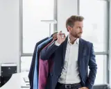 Ein Mann mit Bart trägt einen blauen Anzug und hält mehrere Anzüge zur Reinigung auf einem Bügel.