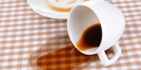 Um Kaffeeflecken zu entfernen, ist hier ein umgekippter Becher mit Kaffee auf kariertem Stoff.