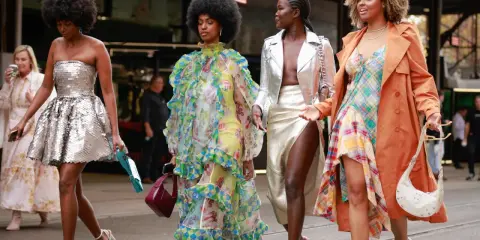 Vier Frauen in auffälligen Outfits. Eine trägt eine silberne Paillettenjacke zum Kleid, eine andere ein buntes Rüschenkleid.
