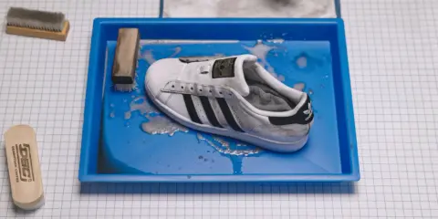 Ein schmutziger weißer Adidas Superstar Schuh liegt in einer blauen Wanne mit Wasser und Seife. Reinigungswerkzeuge liegen daneben.