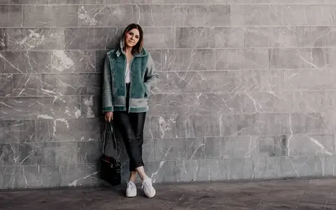 Frau in mintgrüner Lammfelljacke und Lederhose, die weiße Sneakers trägt. Sie steht vor einer grauen Marmorwand.