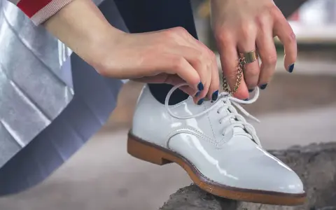 Hände mit dunkelblauem Nagellack binden weiße Lackschuhe. Die Pflege von Lackschuhen ist wichtig, damit sie glänzen.