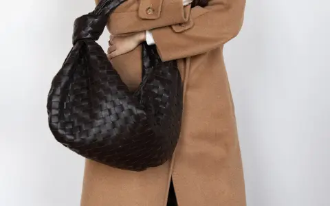 Dunkle, gewebte Tasche mit Knoten-Detail, getragen zu einem Kamelhaarmantel. Sieht aus wie eine Bottega Veneta, aber es ist ein Bottega Veneta Fake.