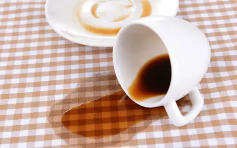Um Kaffeeflecken zu entfernen, ist hier ein umgekippter Becher mit Kaffee auf kariertem Stoff.