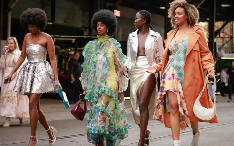 Vier Frauen in auffälligen Outfits. Eine trägt eine silberne Paillettenjacke zum Kleid, eine andere ein buntes Rüschenkleid.
