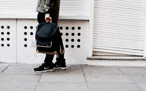 Person mit schwarzem Rucksack und **air max fake** Schuhen steht auf einem Gehweg vor einer weißen Wand mit runden Öffnungen.