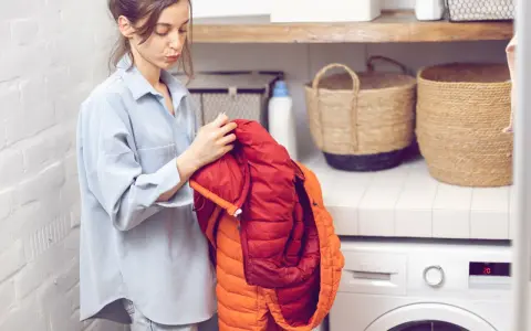 Frau prüft einen roten und orangen Daunenmantel, bereit zum wollmantel waschen. Daneben stehen Wäschekörbe und eine Waschmaschine.