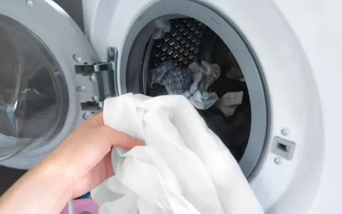 Eine Hand hält ein weißes Hemd, bereit, es in die Waschmaschine zu legen. Die Trommel ist bereits mit anderer Wäsche gefüllt.