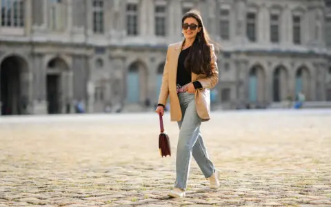 Eine Frau mit langen Haaren und Sonnenbrille spaziert lässig in weiten Jeans und einem beigen Blazer vor einer historischen Fassade.