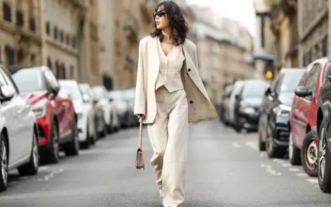 Eine Frau in einem eleganten beigen Hosenanzug mit passendem Westchen und Blazer spaziert durch eine Pariser Straße. Die weiten Hosen und die lässige Silhouette der Schuhe zu Hosenanzug Damen sind perfekt für einen stilvollen Auftritt.