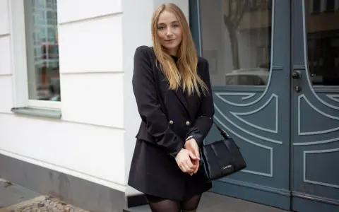 Frau mit blonden Haaren, die ein schwarzes Kleid mit Blazer kombiniert. Sie trägt eine schwarze Tasche und graue Stiefel.