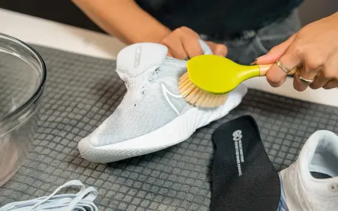 Hände reinigen weiße Mesh-Schuhe mit einer Bürste. Daneben liegen Schnürsenkel und eine Einlegesohle.