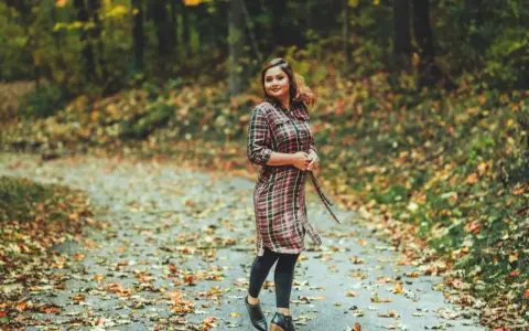 Frau im karierten Kleid mit Leggings steht auf einem herbstlich bedeckten Weg.