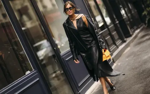 Stilvolle Frau in einem schwarzen Lederkleid und passender Lederjacke, die zeigt, wie man ein schwarzes Kleid kombinieren kann. Orange Handtasche als Farbtupfer.