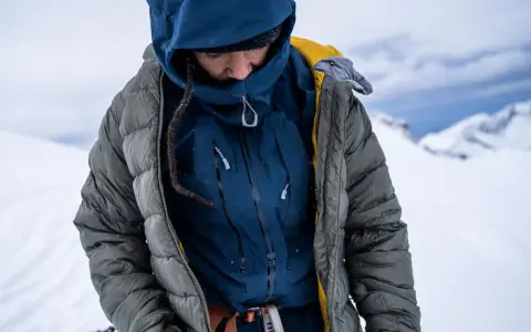 Person in Daunenjacke im Schnee. Die Jacke ist grau, darunter eine blaue Schicht. Perfekt für das Daunenjacke waschen nach der Tour.