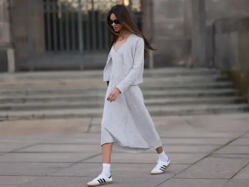 Frau in grauem Strick-Abendkleid mit Cardigan und weißen Sneakern, die lässig über einen Platz geht.