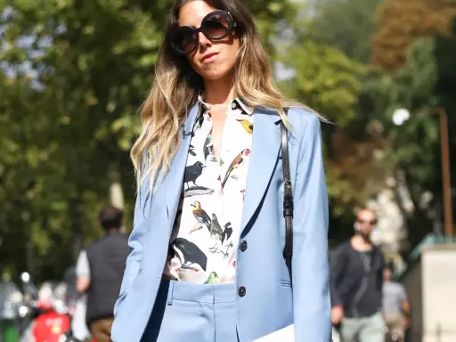 Eine Frau kombiniert einen blauen Blazer mit einem Hemd mit Vogelmotiv und auffälliger Sonnenbrille.