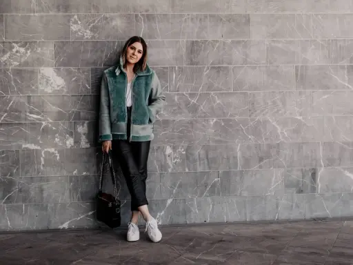Frau in mintgrüner Lammfelljacke und Lederhose, die weiße Sneakers trägt. Sie steht vor einer grauen Marmorwand.