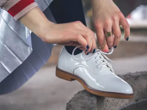 Hände mit dunkelblauem Nagellack binden weiße Lackschuhe. Die Pflege von Lackschuhen ist wichtig, damit sie glänzen.